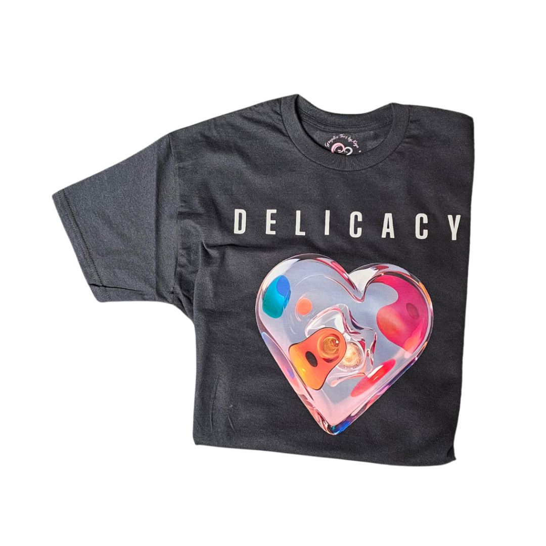 A black T-shirt with the word DELICACY in white capital letters above a large, colorful, translucent heart graphic on the front.