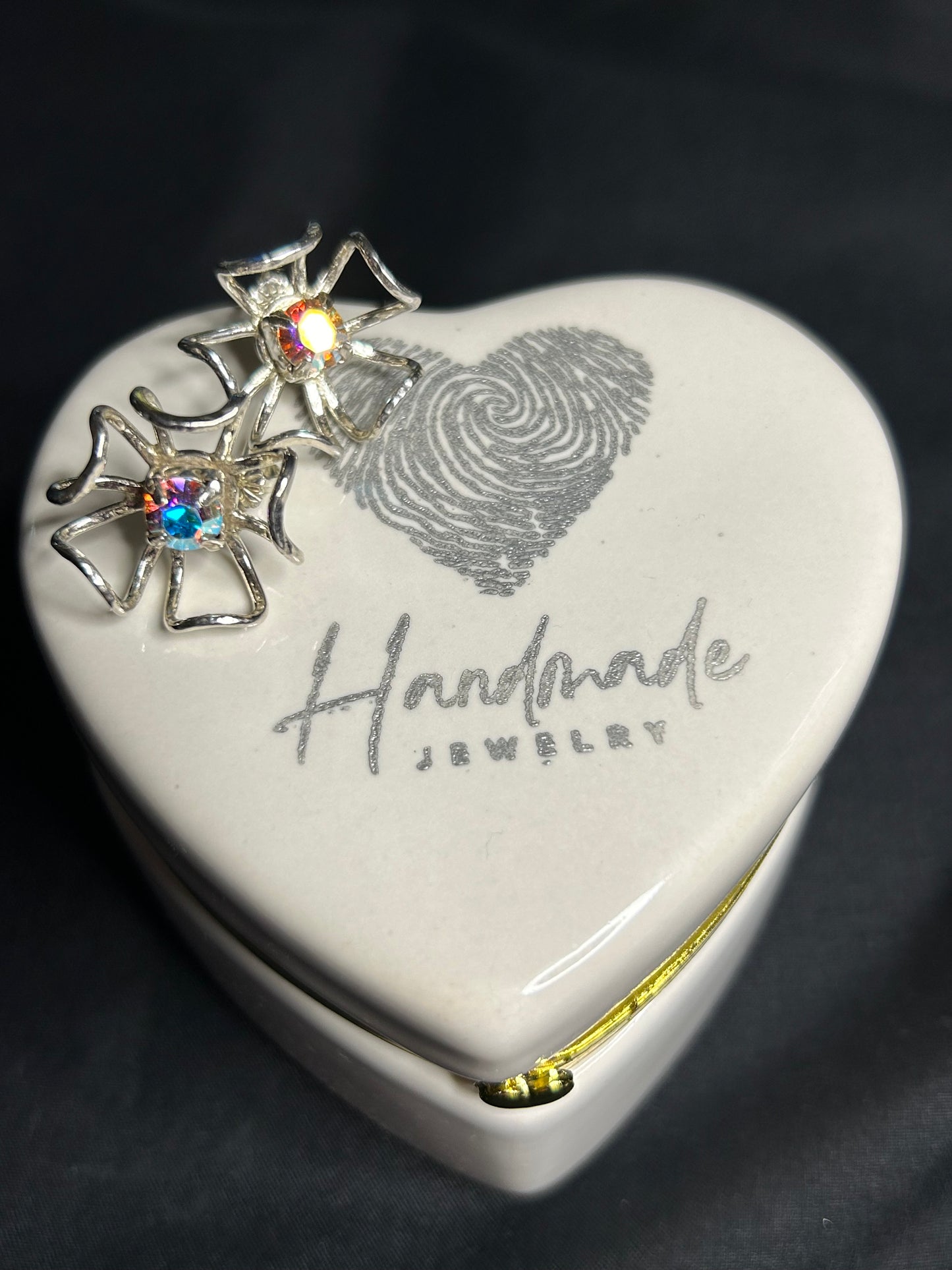 A heart-shaped ceramic jewelry box labeled Handmade Jewelry with a fingerprint design holds two Stunning Handcrafted Sterling Silver Flower Earrings w AB Prism Crystal by Delseys Treasures on its lid, set against a black background.
