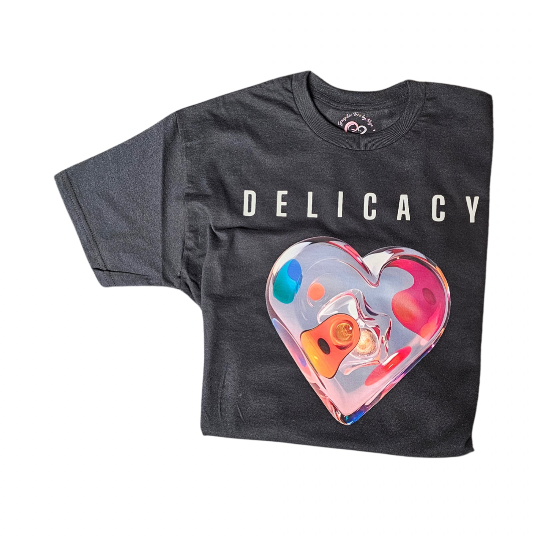 The Try My Closet Delicacy Heart Color Purple Changing Graphic Tee features “DELICACY” in white above a large, colorful abstract heart graphic on the front. The tees color changes to purple in UV light.
