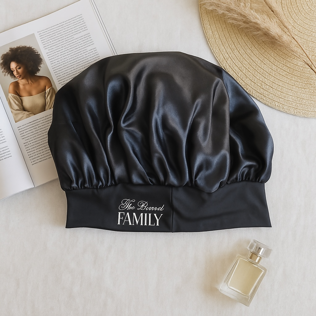 A black satin bonnet with “The Bonnet FAMILY” embroidered in white sits on a white surface next to a beige hat, perfume bottle, dried grass, and an open magazine showing a woman.