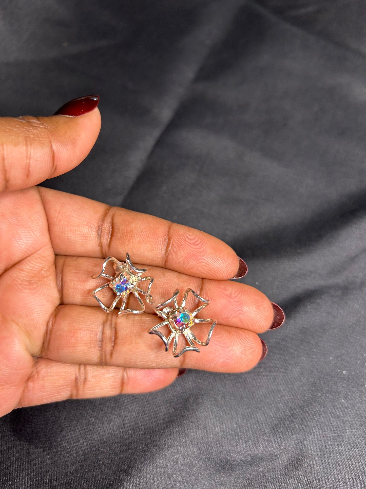 A hand with painted nails holds two Delseys Treasures Stunning Handcrafted Sterling Silver Flower Earrings, each featuring a vibrant AB prism crystal at the center, against a dark background.