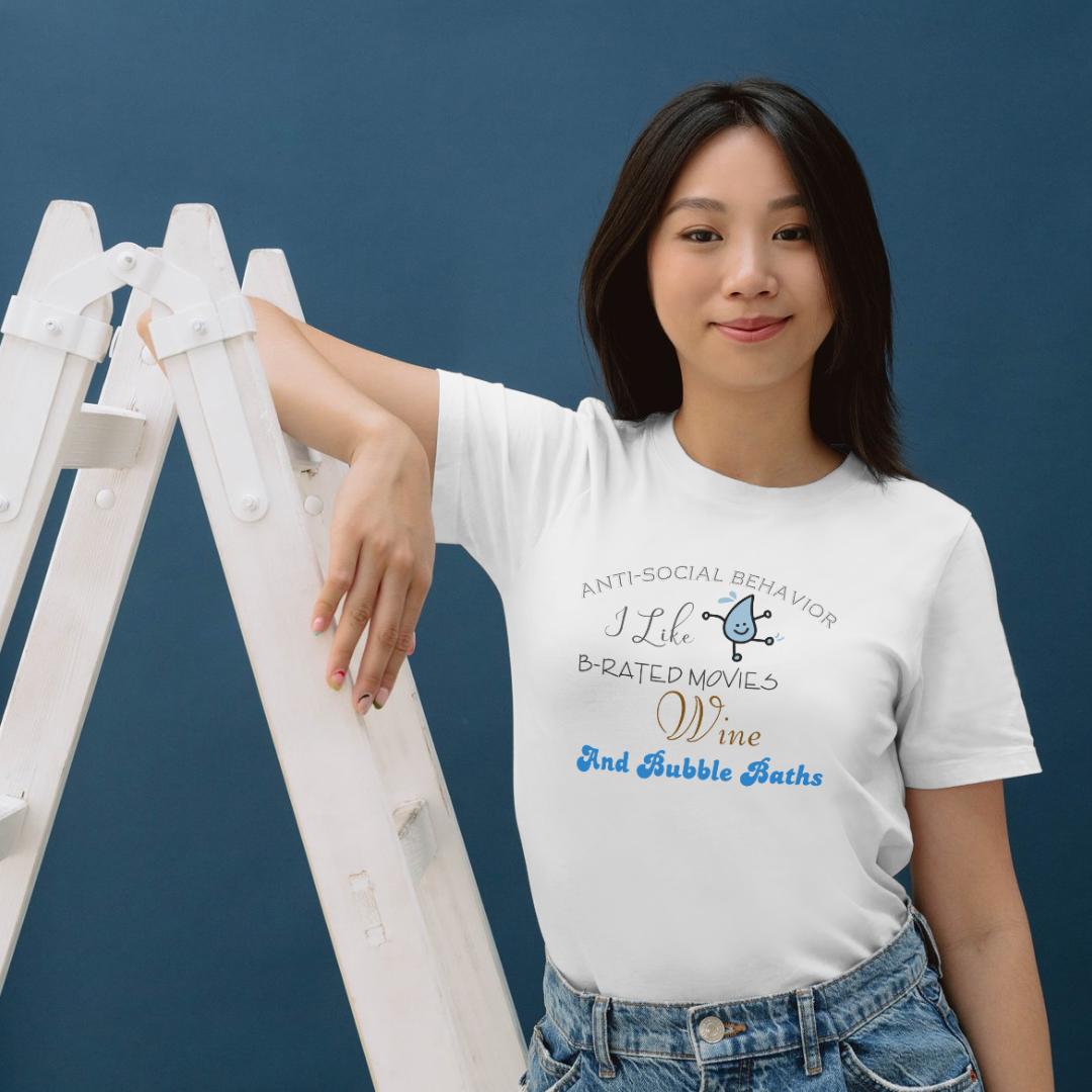 A woman wearing the Try My Closet Anti-social Behavior Graphic Tee stands smiling by a white ladder against a blue background. The white cotton tee features playful text and custom orders are available.