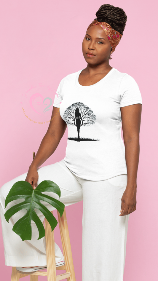 A woman in white pants and a Shes Rooted in Family Graphic Tee by Try My Closet sits on a stool with a green monstera leaf, wearing a patterned headband against a pink background—embodying the spirit of being rooted in family.